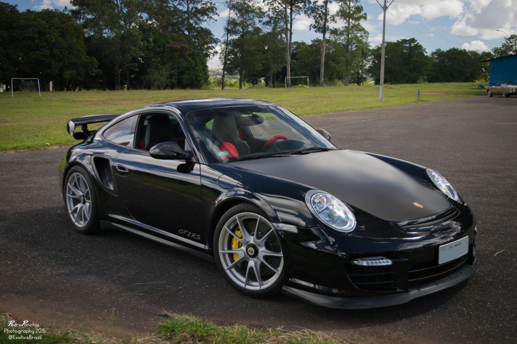 Porsche 911 GT2 RS da geração 997, preto com capô de fibra de carbono e pinças amarelas, estacionado num terreno gramado em Brasília